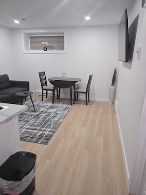 Un salon moderne comprenant un canapé, une table à manger avec des chaises, un petit tapis, un téléviseur fixé au mur et une fenêtre avec des stores.
