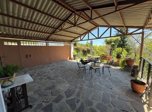 Um pátio externo com piso de pedra, área de estar coberta com cadeiras e uma mesa, e plantas em vasos.
