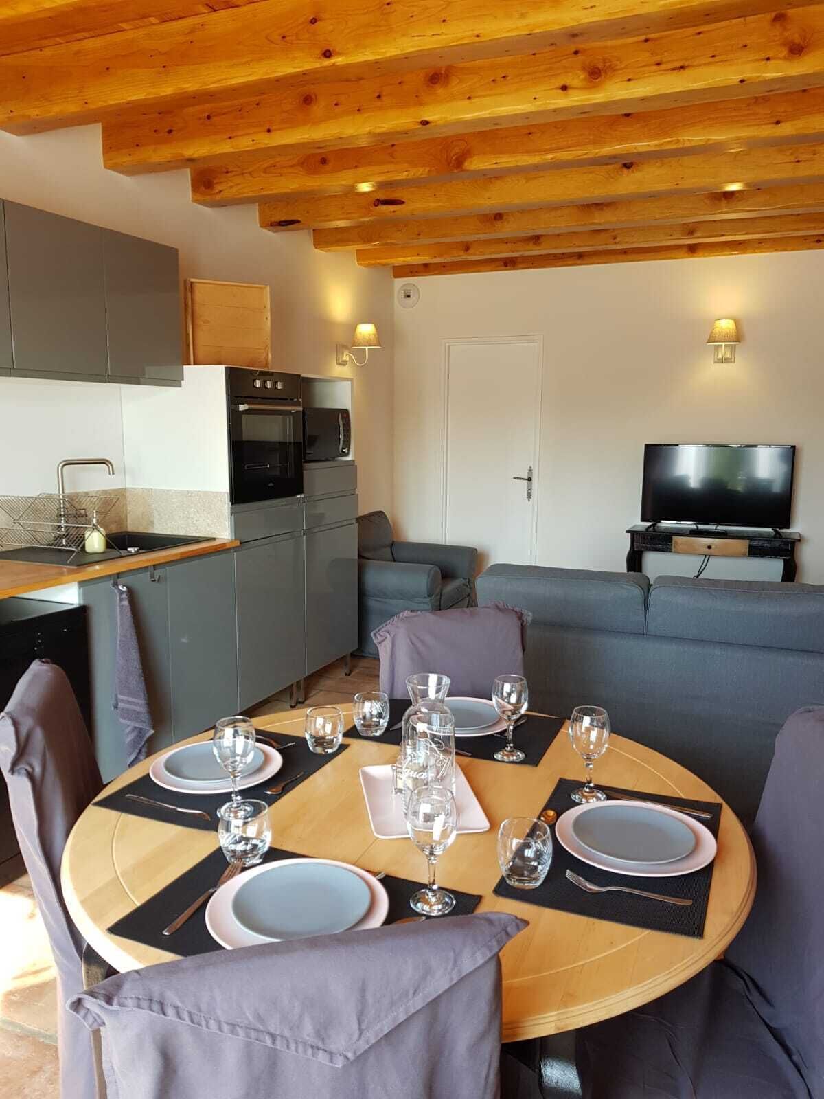 A modern kitchen with a built-in oven and microwave, a dining area with a round table set for four, and a living area with a sofa and a flat-screen TV.