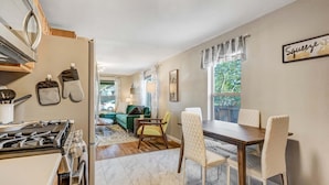 A kitchen with a stove, oven, and cabinets, a dining area with a table and chairs, and a living room with a sofa and a window. - 1014a Swope (Colorado Springs)