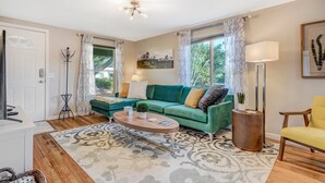 A living room with a green sofa, a round coffee table, and a yellow armchair. There is a large window with curtains, a floor lamp, and a painting on the wall. - 1014a Swope (Colorado Springs)