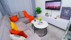 En moderne stue med en grå sofa, orange og gule puder, et hvidt sofabord, et fjernsyn på væggen og et plakat med en løve.