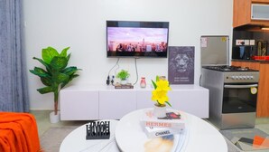 Ein modernes Wohnzimmer mit Flachbildfernseher, einer weißen Unterhaltungsanlage, einer Küchenzeile mit Herd und einer großen Zimmerpflanze.