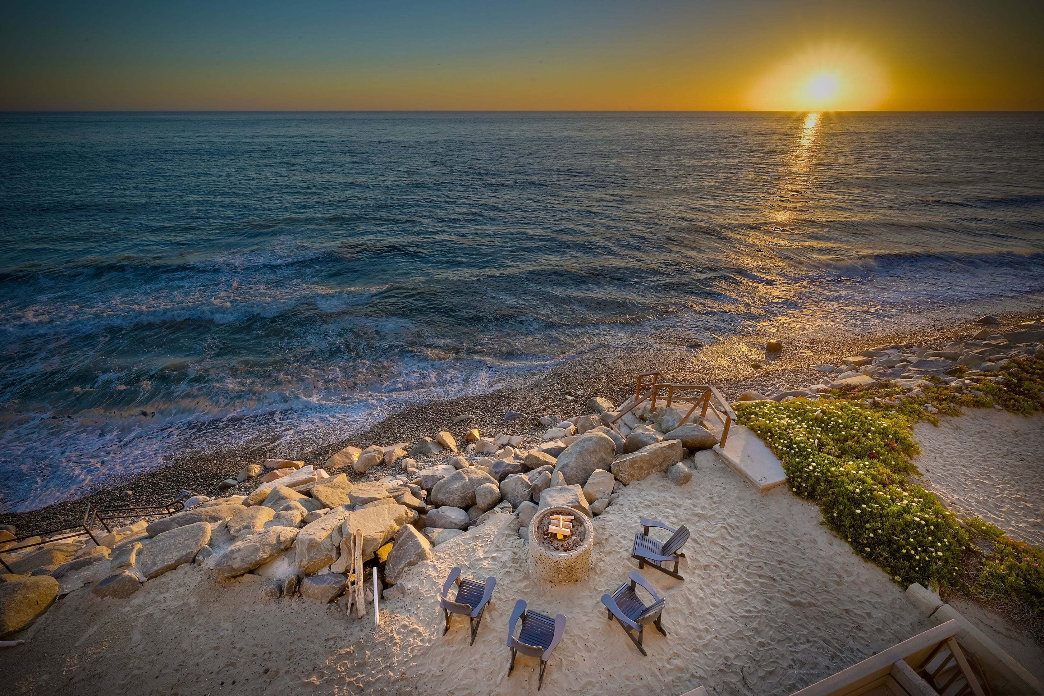 A beachside seating area with a fire pit, chairs, and a view of the ocean at sunset.
