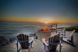 Paisaje playero con una fogata, sillas Adirondack y vista al atardecer sobre el océano.