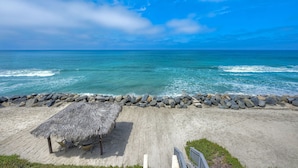 Ein Strand mit einer strohgedeckten Struktur, Stühlen, einem Betonweg und einer felsigen Küstenlinie.