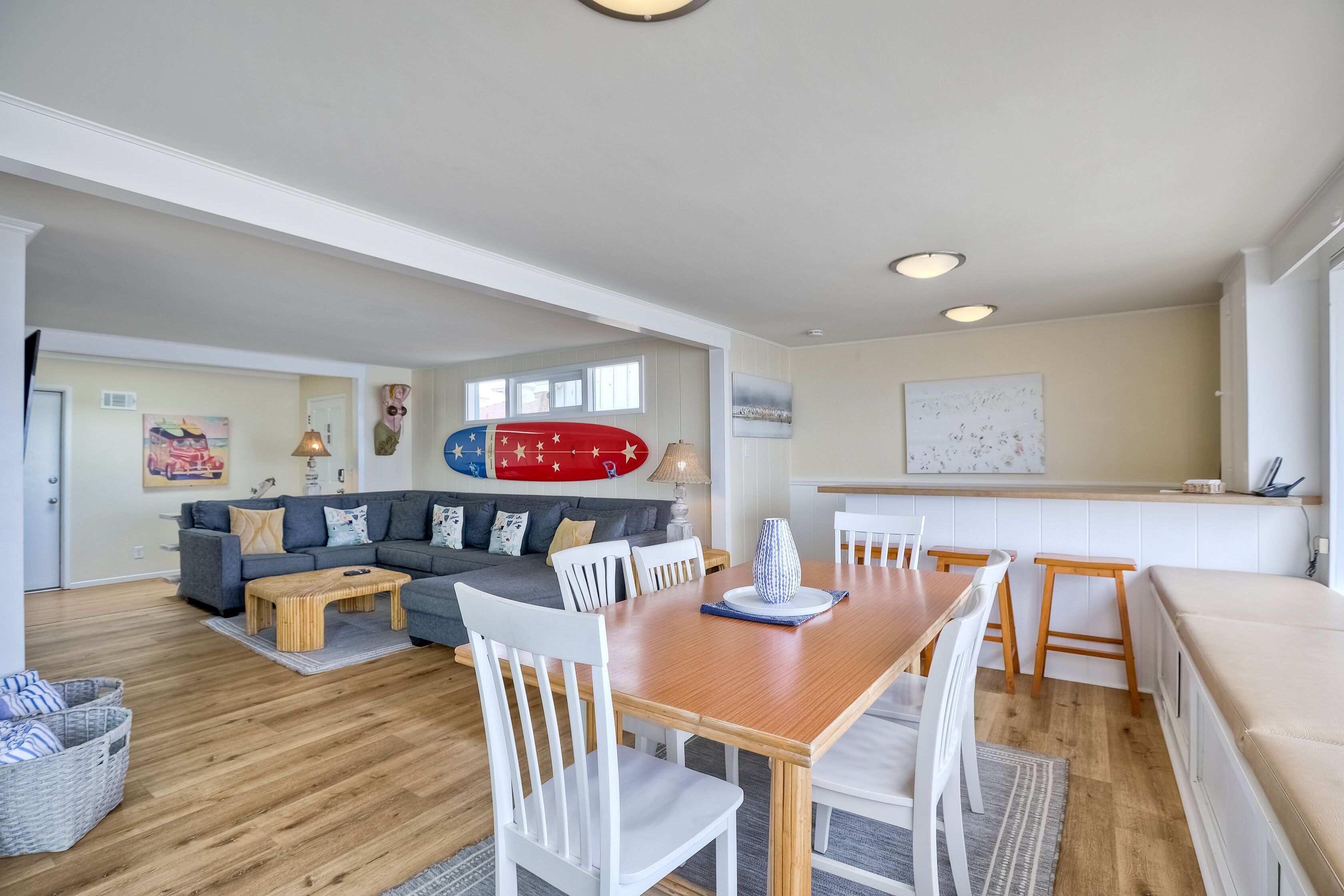 Amplio espacio habitable con una mesa de comedor de madera, sillas blancas, un sofá gris y una decoración de tabla de surf roja.