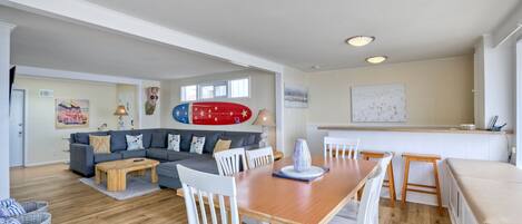 Amplio espacio habitable con una mesa de comedor de madera, sillas blancas, un sofá gris y una decoración de tabla de surf roja.