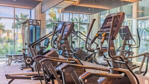 Een sportschool met loopbanden en elliptische trainers, grote ramen en uitzicht op palmbomen.