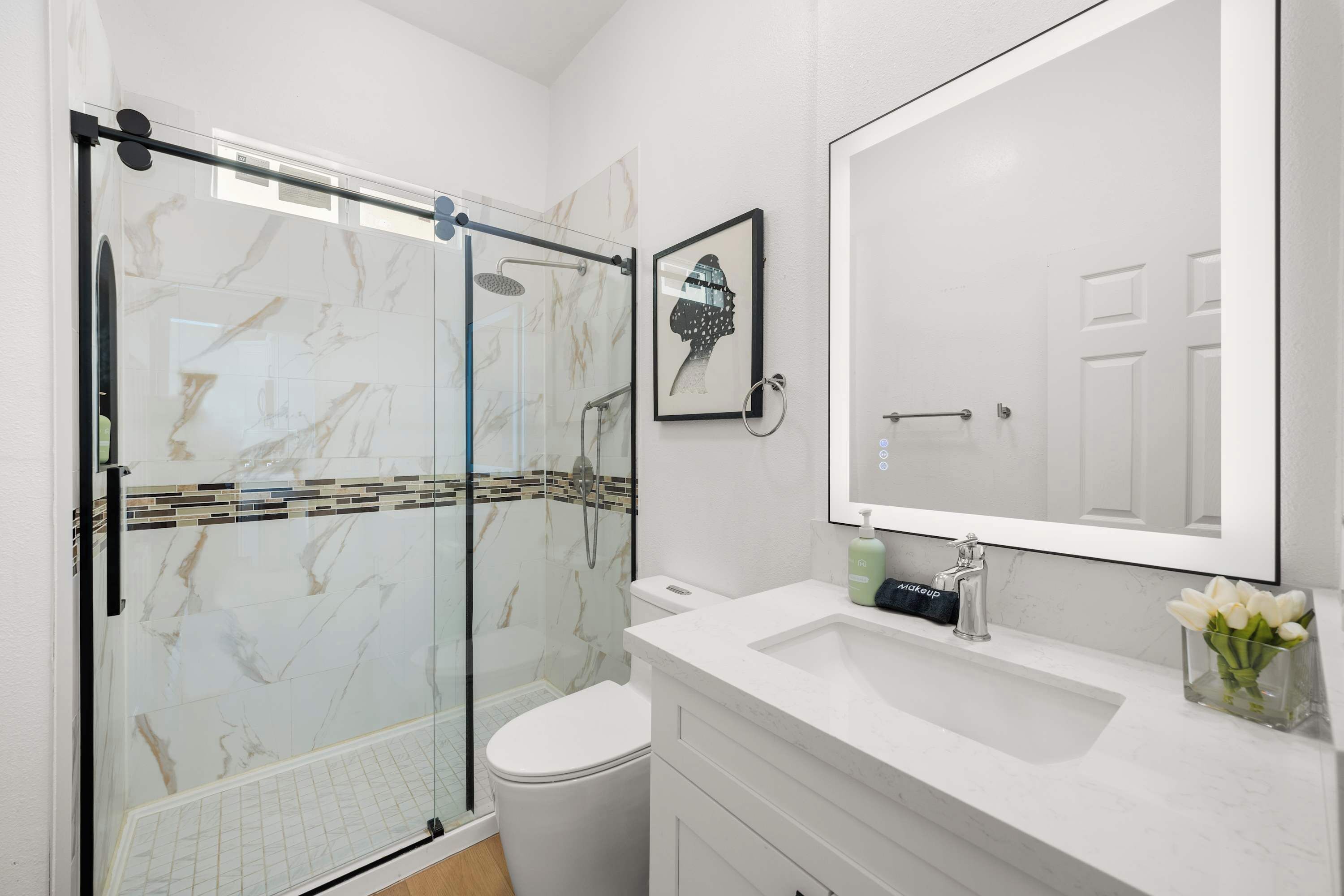 Un baño moderno con ducha envuelta en vidrio, inodoro blanco, espejo grande y lavamanos blanco con un florero.