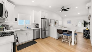A modern kitchen with white cabinets, stainless steel appliances, a dining area with a white table and bar stools, and a ceiling fan.