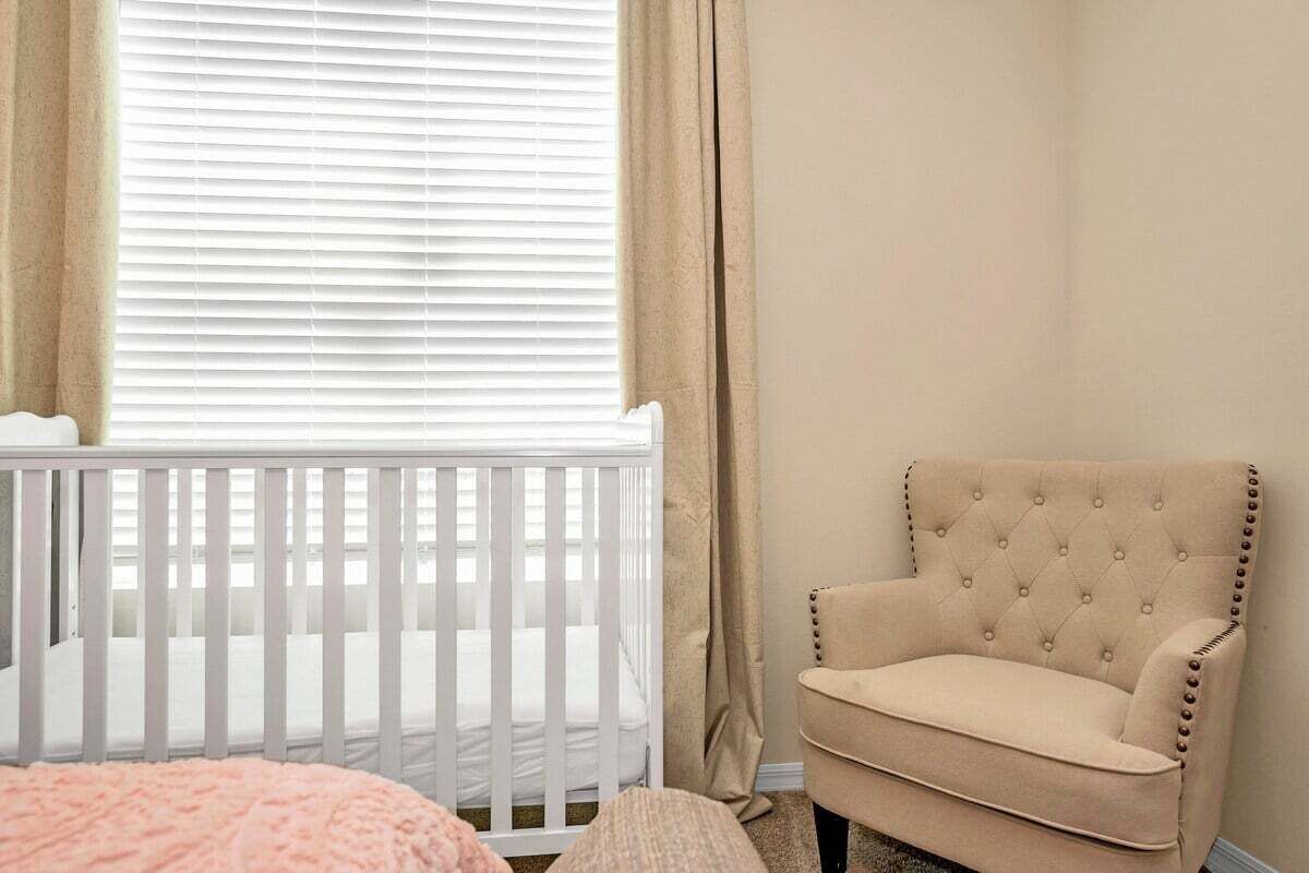 Une pièce avec un berceau blanc, un fauteuil beige et une fenêtre avec des stores.