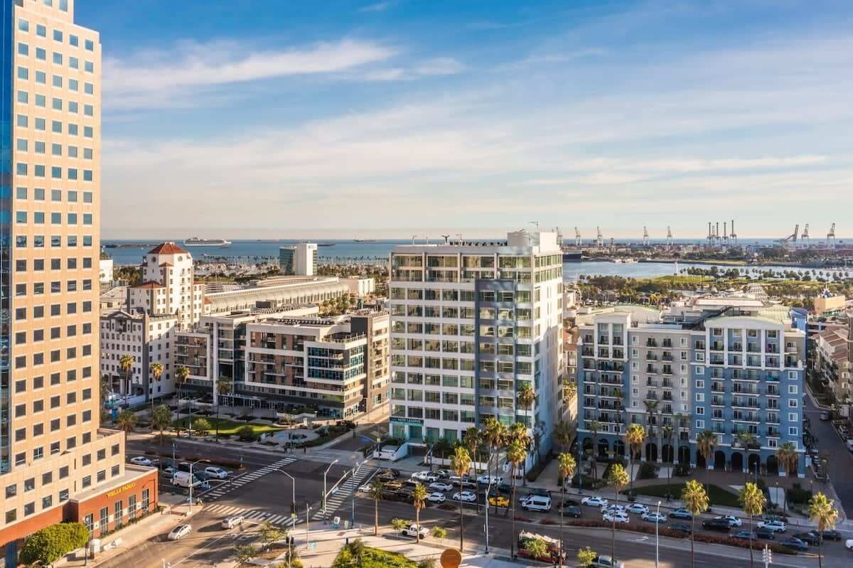 A high-rise hotel with multiple floors, a parking lot, and a view of the sea and industrial area in the background.