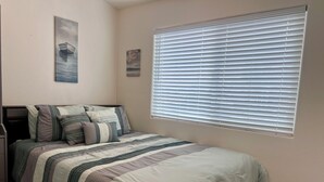 Een slaapkamer met een bed, kussens, een raam met zonwering en kunst aan de muur.