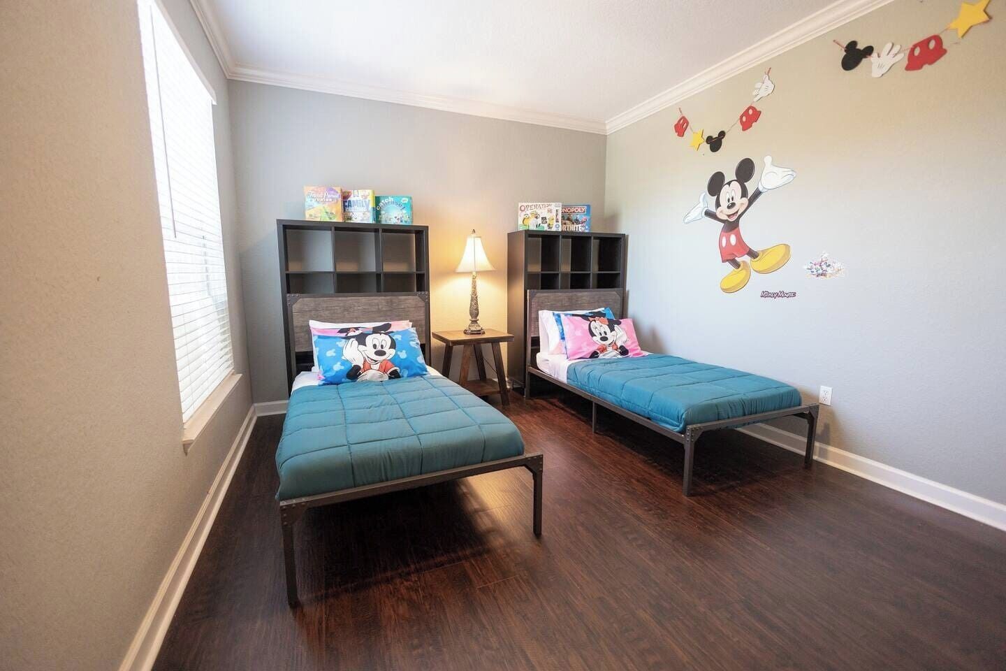 Ein Kinderzimmer mit zwei Betten, jedes mit einem Mickey Mouse Kissen, einem Bücherregal und einer Wandmalerei von Mickey Mouse.