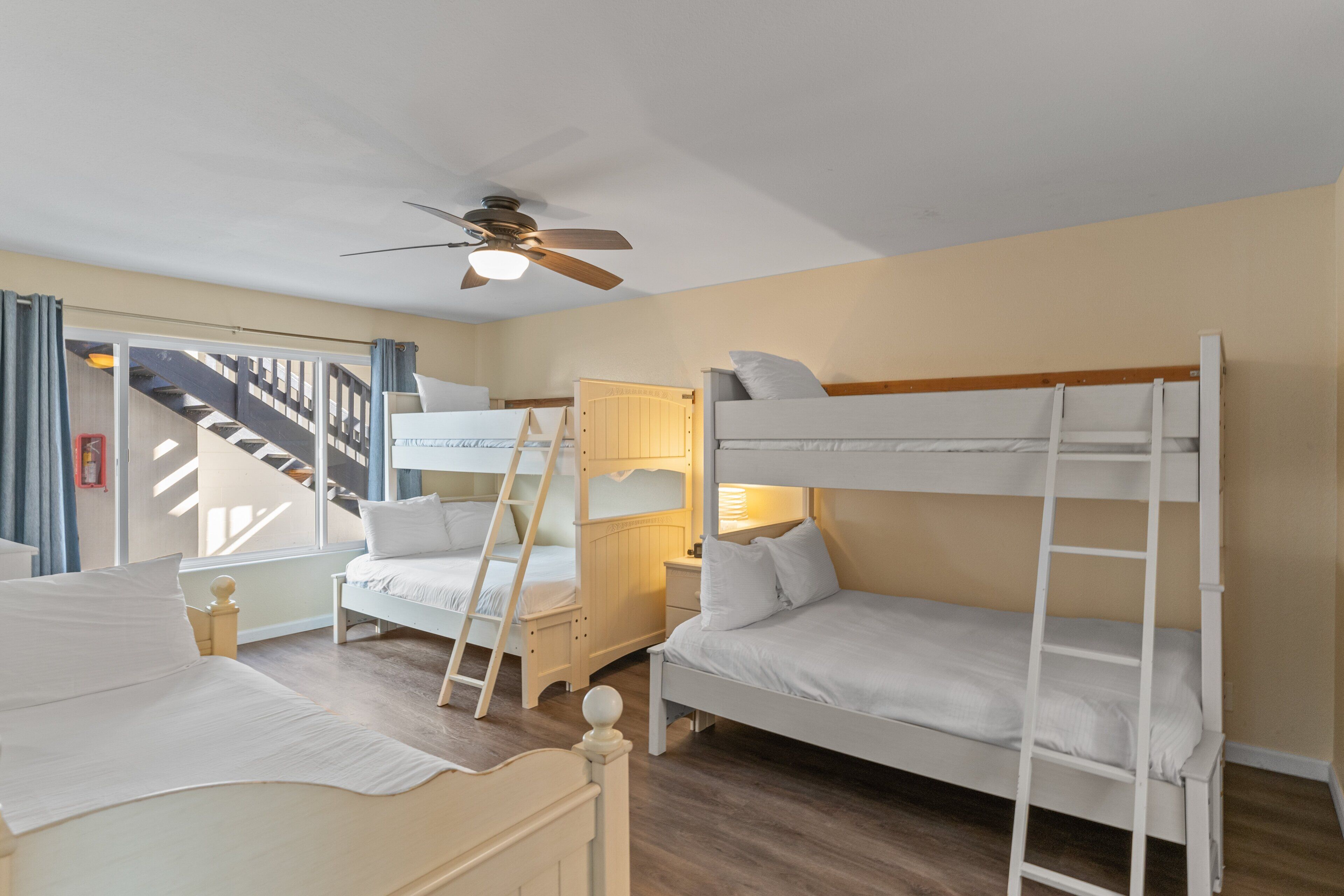 A room with bunk beds, a ceiling fan, a window with curtains, and a staircase.