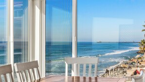 A view from a balcony overlooking a beach with a wooden table and white chairs, a pier in the distance, and a clear sky. - 1317 S Pacific - Entire Home (Oceanside)