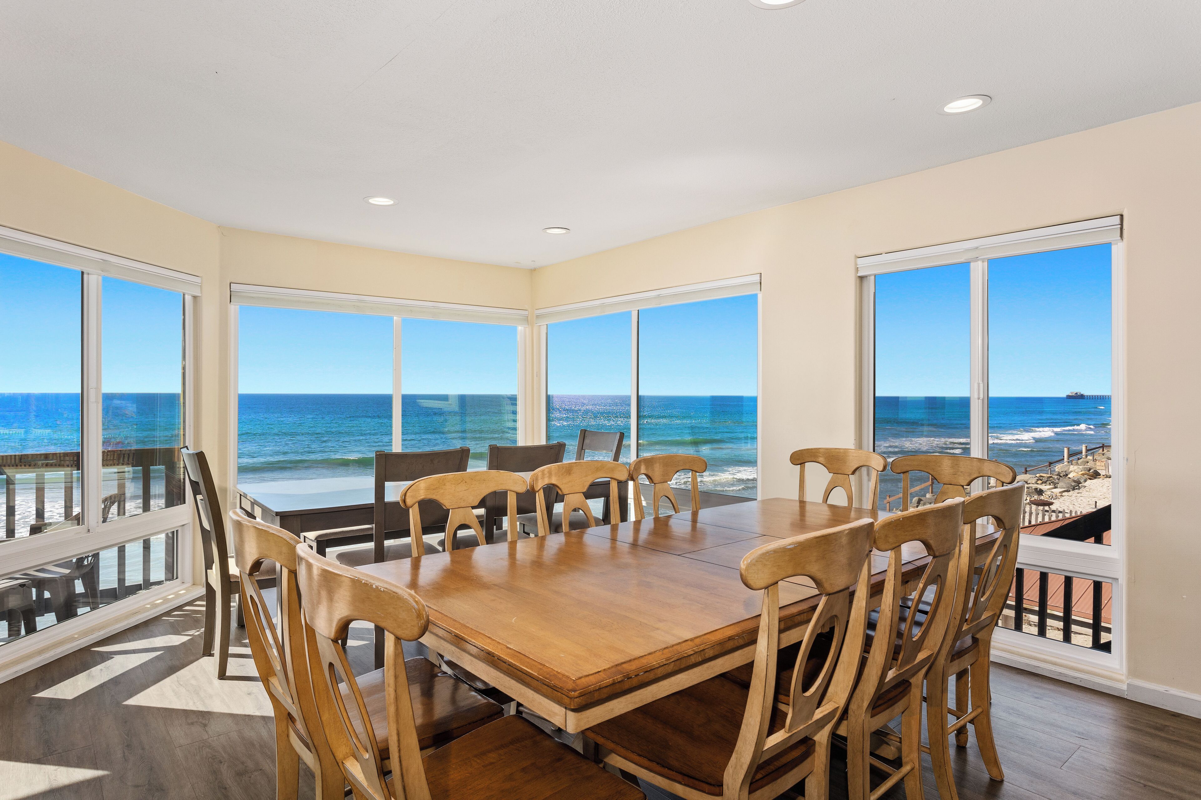 A dining area with a large table and chairs, large windows with ocean views, and a balcony with a railing.
