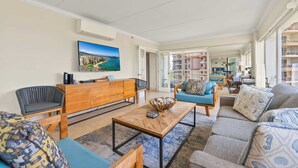 A spacious living room with a large flat-screen TV, comfortable seating, and a view of the city through large windows. - @ Marbella Lane - Cozy 2BR w/ Amazing Ocean Views (Honolulu)