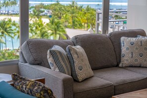 A grey sofa with patterned cushions, a view of a beach with palm trees, and a balcony railing. - @ Marbella Lane - Cozy 2BR w/ Amazing Ocean Views (Honolulu)