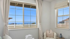 Une chambre d’hôtel avec un lit, une chaise et une petite table. Il y a deux fenêtres avec des rideaux, et la vue donne sur la mer et les bâtiments.