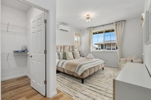 Ein Schlafzimmer mit einem großen Bett, einem Nachttisch, einer Kommode, einem Fenster mit Vorhängen und einer Klimaanlage.