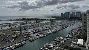 A marina with numerous boats docked, a road with vehicles, and a city skyline in the background. - @ Marbella Lane Dreamy Sunset Views by the Sea (Honolulu)