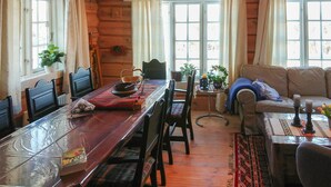 L’intérieur d’un chalet confortable, avec des murs en bois, une table à manger, des chaises, un canapé et de grandes fenêtres ornées de rideaux.