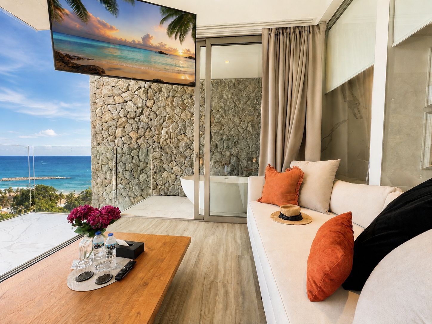 Una sala de estar moderna con una pared de piedra, un sofá blanco con cojines anaranjados, una mesa de centro con un jarrón de flores y una pantalla de televisión grande mostrando una escena de playa.