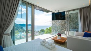 A hotel room with a large bed, a sofa, a TV mounted on a stone wall, and a view of the sea and mountains. - Sea Theatre Kata (Karon)