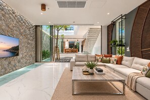 A modern living room with a stone wall, a large flat-screen TV, a sofa set, a coffee table, and a staircase leading to an upper level. - Sea Theatre Kata (Karon)