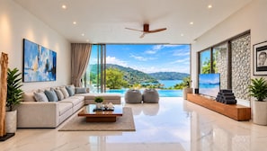 Un salon moderne avec un canapé, une table basse et un téléviseur. Une grande fenêtre offre une vue sur une plage et des collines.