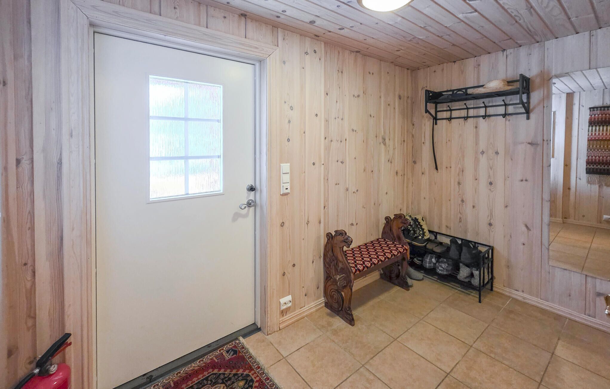 Una habitación con paredes de madera, una puerta blanca, un banco con cojín estampado y un zapatero.