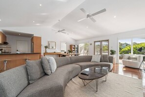 A spacious living area with a large gray sectional sofa, a glass coffee table, and a modern kitchen in the background. - The Coastal Garden House - With Spa! (Rye)