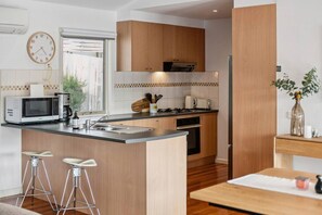 A modern kitchen with wooden cabinets, a built-in microwave, a sink, and a stove. There are two bar stools and a dining table with a vase of flowers. - The Coastal Garden House - With Spa! (Rye)