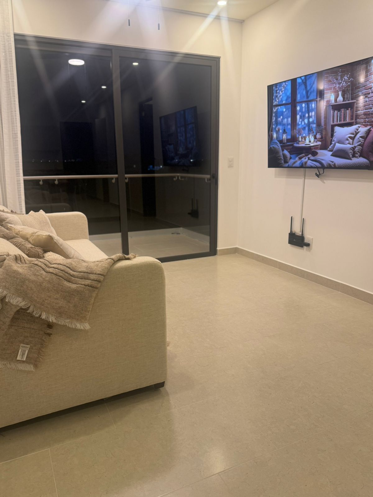 Un salon avec un canapé beige, une porte vitrée donnant sur un balcon et un téléviseur à écran plat fixé au mur.