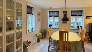 Een eetruimte met een ronde tafel, houten stoelen en een kroonluchter. Er zijn ramen met jaloezieën, een boekenkast en potplanten.