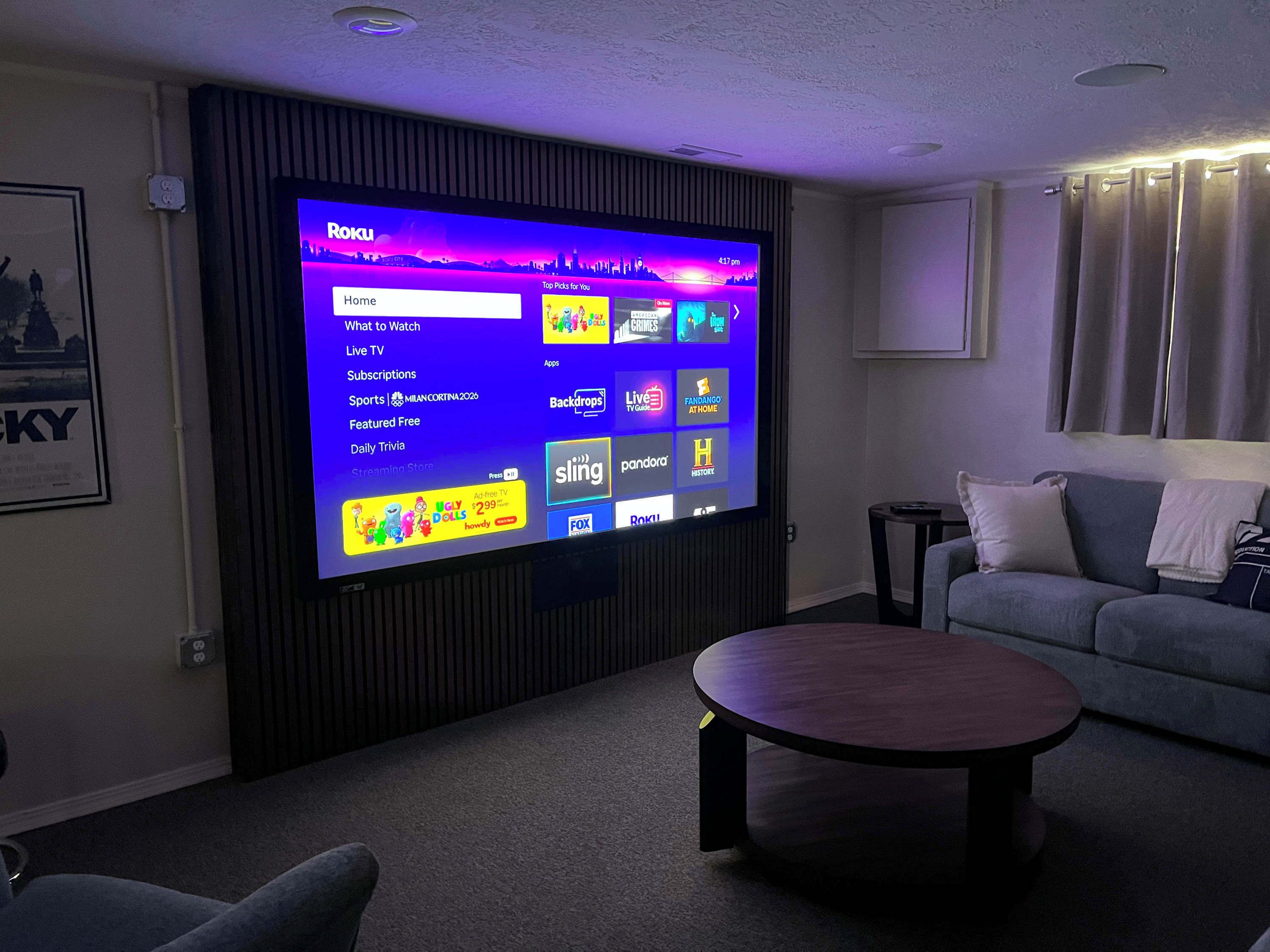 Una sala de estar con un televisor de pantalla plana grande montado en la pared, un sofá gris, una mesita de centro redonda y un cuadro enmarcado en la pared.