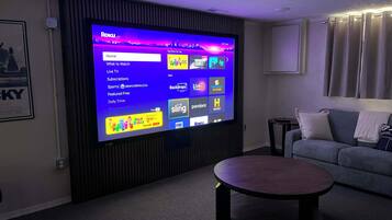 Una sala de estar con un televisor de pantalla plana grande montado en la pared, un sofá gris, una mesita de centro redonda y un cuadro enmarcado en la pared.