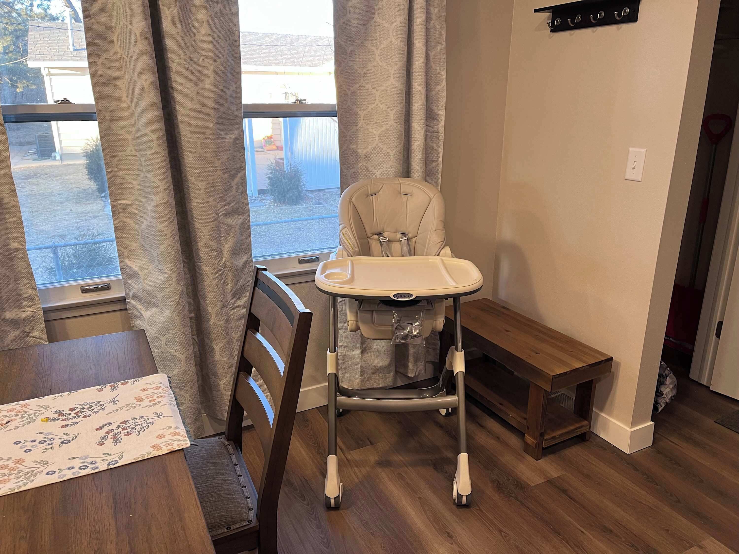 Una habitación con una mesa de comedor de madera, una trona con bandeja, un banco y una ventana con cortinas.