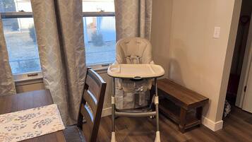 Una habitación con una mesa de comedor de madera, una trona con bandeja, un banco y una ventana con cortinas.