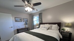A bedroom with a bed, a nightstand, a lamp, a ceiling fan, a window with curtains, and wall art.
