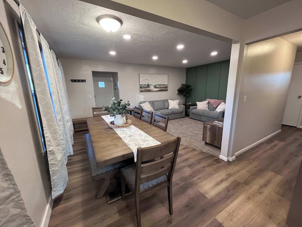 A dining area with a wooden table and chairs, a vase with flowers, and a rug. There is a living area with a couch and a plant. The room has wooden flooring and recessed lighting.