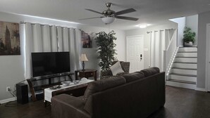 Una sala de estar con una televisión de pantalla plana, un ventilador de techo, un sofá, una mesa de centro y una escalera.