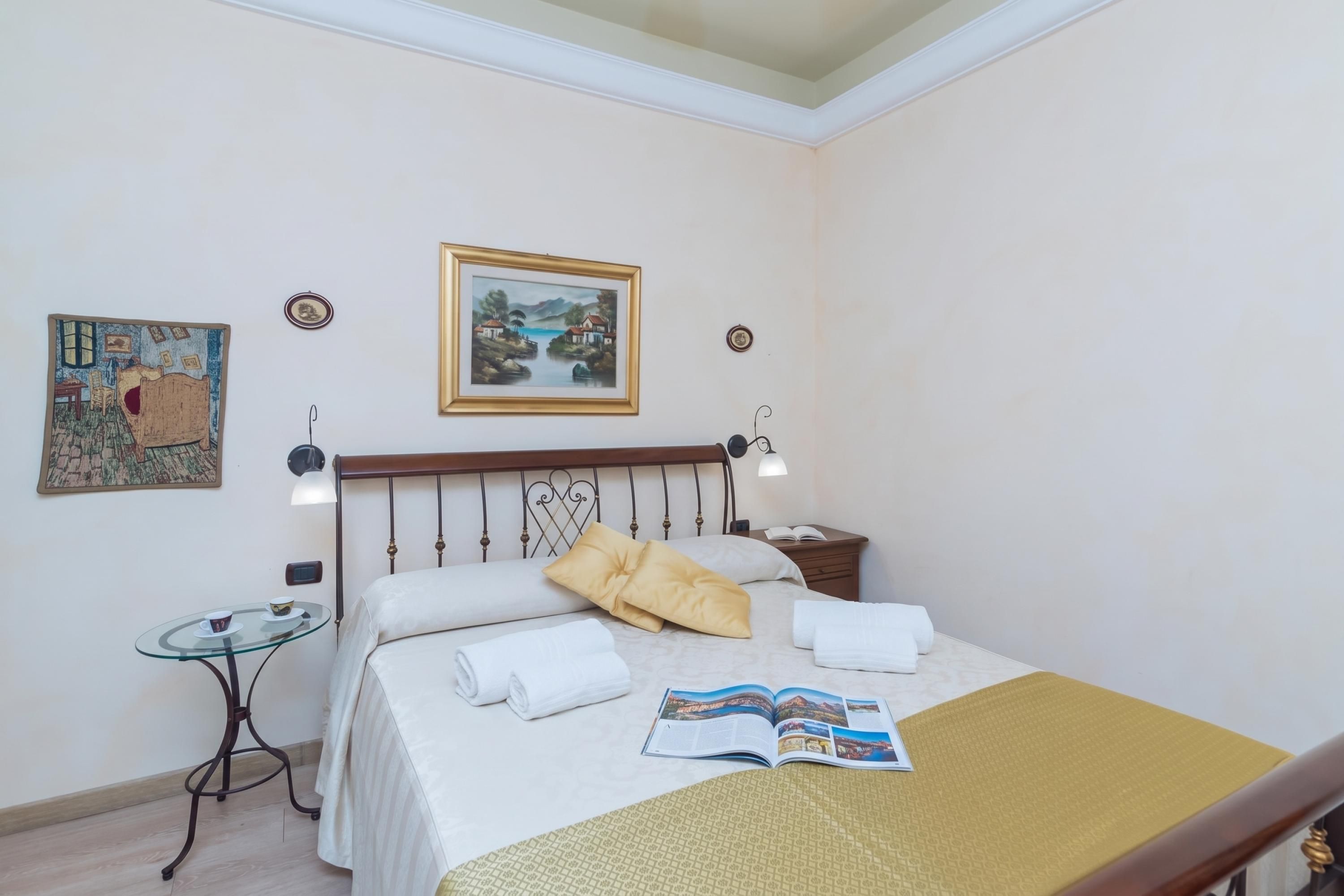 A hotel room with a bed, a bedside table, a reading lamp, a small table with a cup, and a framed painting on the wall.
