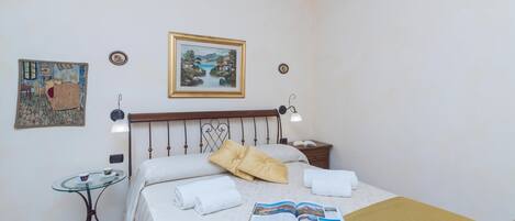 A hotel room with a bed, a bedside table, a reading lamp, a small table with a cup, and a framed painting on the wall.