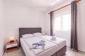 A hotel room with a large bed, a bedside table with a lamp, a window with curtains, and a magazine on the bed. - Pool Apt 4 - Novi Vinodolski (Ground), Novi Vinodolski, Croatia (Novi Vinodolski)