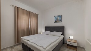 A bedroom with a bed, a nightstand with a lamp, a painting on the wall, and a window with curtains. - Pool Apt 3 - Novi Vinodolski (Ground), Novi Vinodolski, Croatia (Novi Vinodolski)