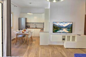 A modern kitchen with white cabinets, a dining area with a table set for two, and a flat-screen TV mounted on the wall displaying a scenic landscape.
