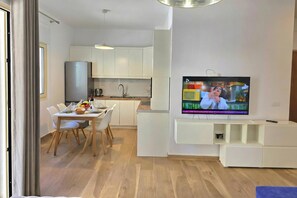 Ein modernes Wohnzimmer mit einem an der Wand montierten Flachbildfernseher, einem Essbereich mit Tisch und Stühlen sowie einer Küche mit weißen Schränken und einem Edelstahl-Kühlschrank.
